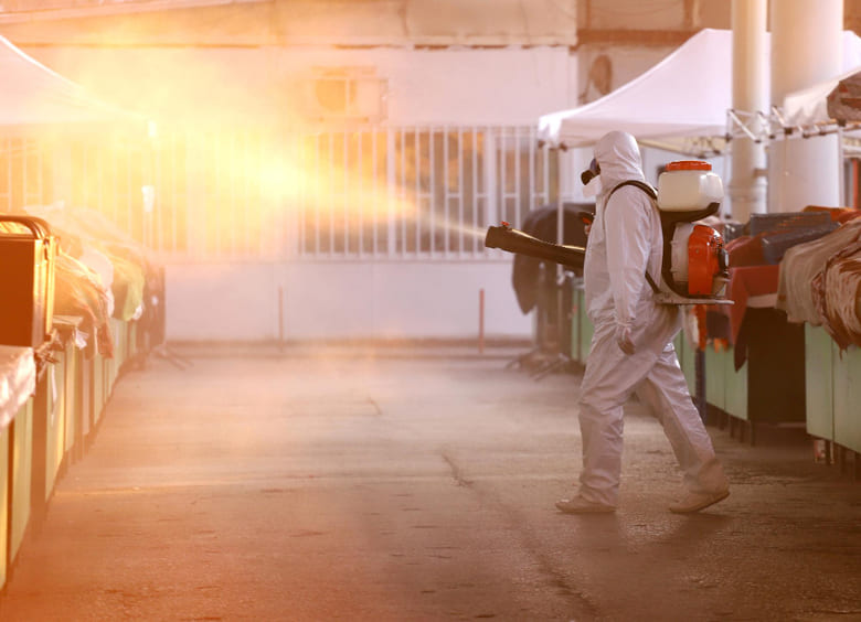 Operatore con DPI protettivi che esegue un intervento di fumigazione in un’area di mercato o magazzino, utilizzando un nebulizzatore per il controllo degli infestanti